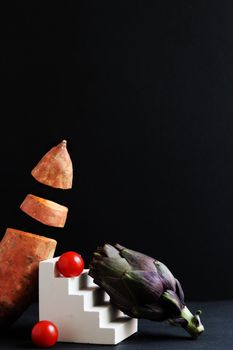 Creative arrangement of vegetables with artichoke, tomatoes, and yams against a dark backdrop.