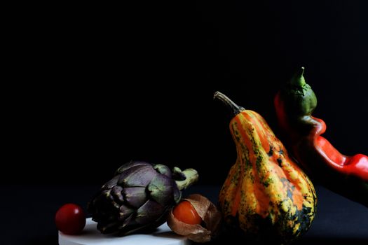 A dramatic still life of artichoke, gourd, and pepper against a black background.