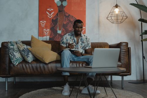 Free Man sitting on sofa in modern living room working on laptop, showcasing a cozy work-from-home setup. Stock Photo