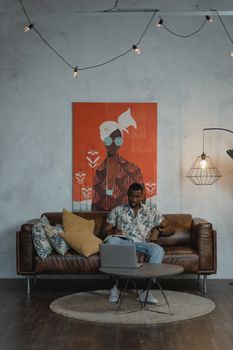 Young man working from home on a laptop, sitting on a sofa in a modern cozy living room.