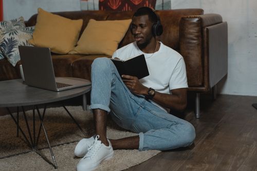 Free Young man sitting on the floor, wearing headphones, taking notes with a laptop nearby in a cozy living room. Stock Photo