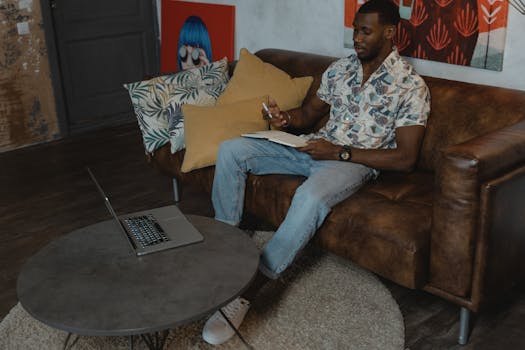 Young man taking online class from home, sitting on a sofa with a laptop and notebook.