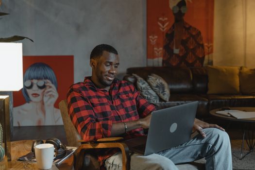 African American man in plaid shirt working from home on laptop, surrounded by art in a cozy room.
