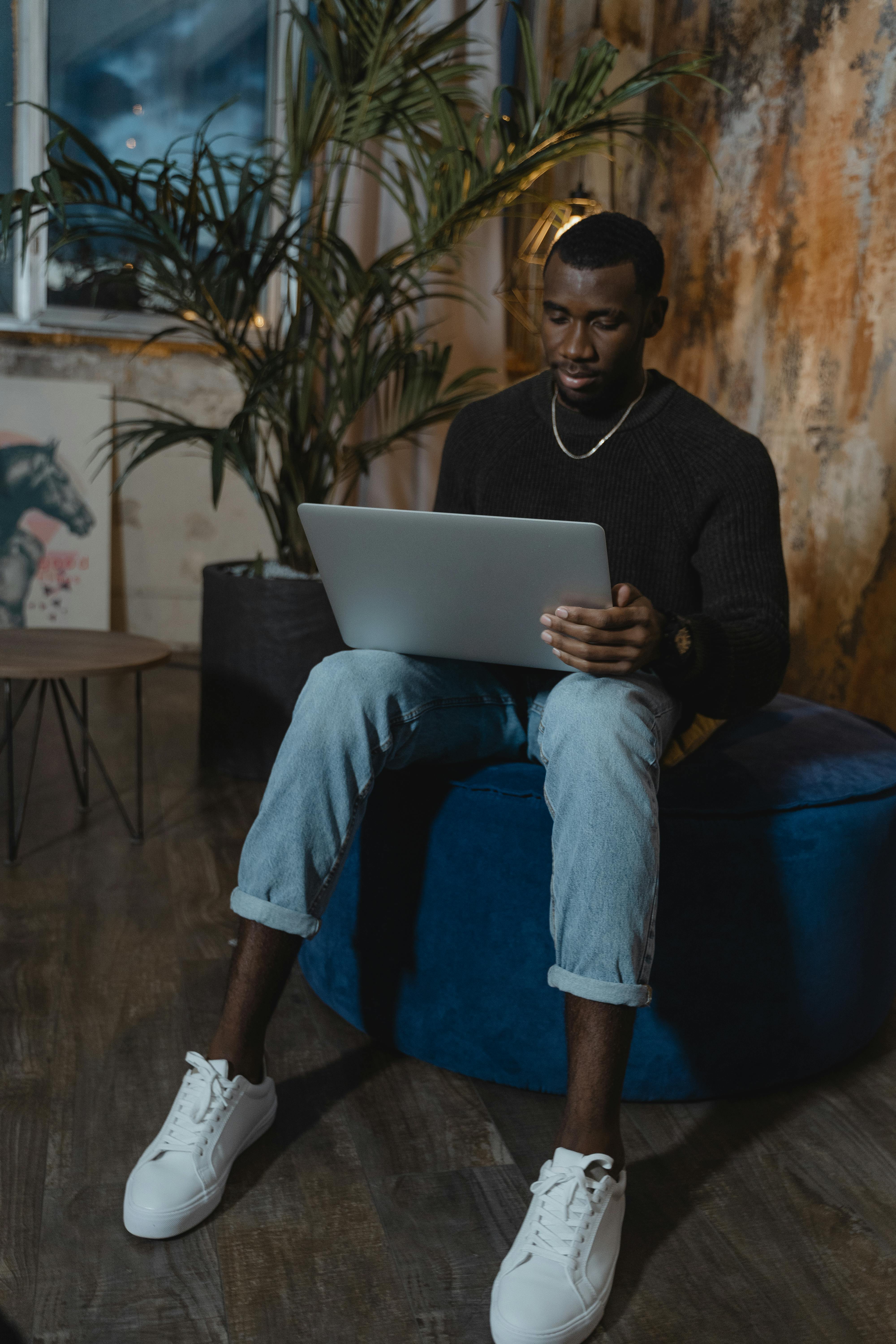 man in black long sleeve shirt and blue denim jeans sitting on blue chair using silver laptop