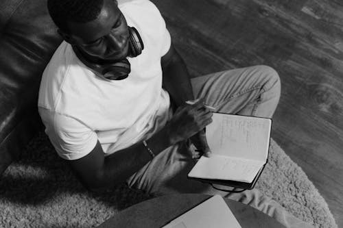 Grayscale Photo of a Man Sitting on the Rug while Holding a Notebook