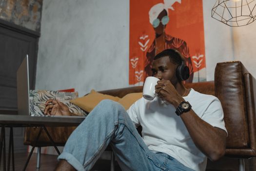Casual young man in headphones enjoying coffee while working remotely with laptop indoors.