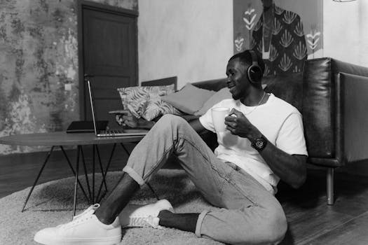 Black and white image of a man with headphones, enjoying coffee and working on a laptop.