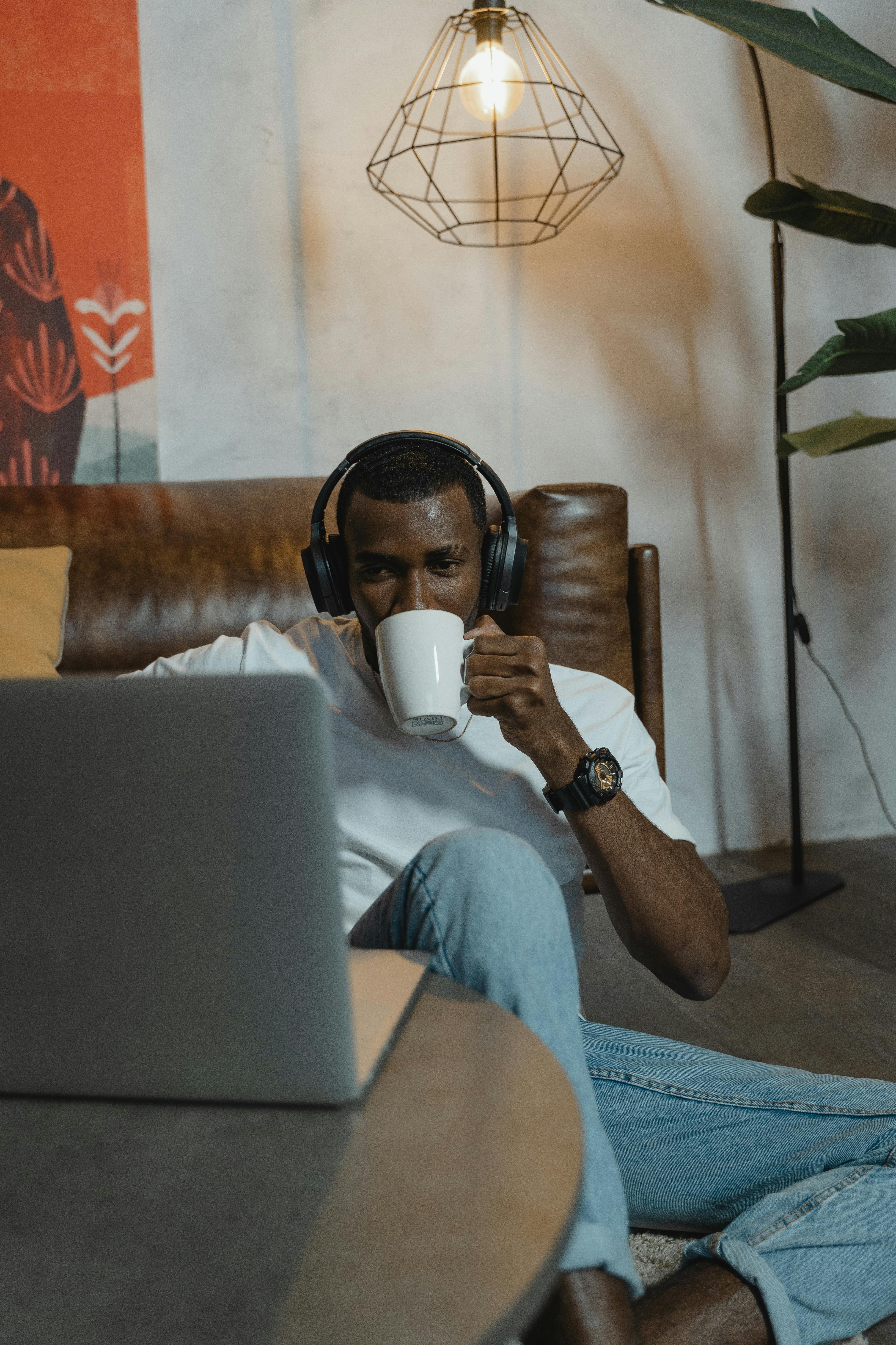 一名男子戴着耳机，舒适地坐在室内，一边喝咖啡一边使用笔记本电脑