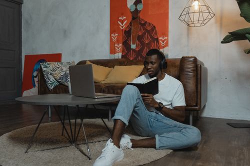 Free A man sitting on the floor in a stylish living room, working remotely on his laptop and writing in a notebook. Stock Photo
