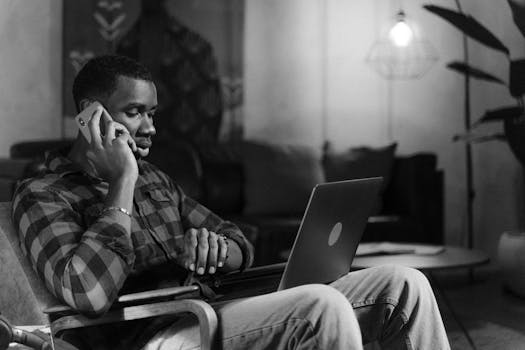 A man in a flannel shirt working remotely with a laptop and phone in a cozy, grayscale setting.