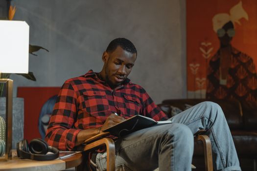 一名穿着格子衬衫的非洲裔美国男子坐在室内，在笔记本上写字
