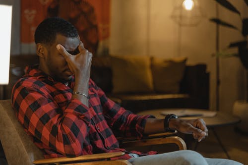 Man in Red Black and White Plaid Dress Shirt Sitting on Brown Wooden Armchair Feeling Exhausted