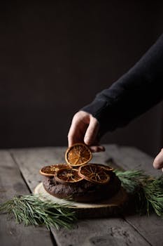 A hand decorates a homemade chocolate cake with dried orange slices on a wooden surface, perfect for cozy winter moments.