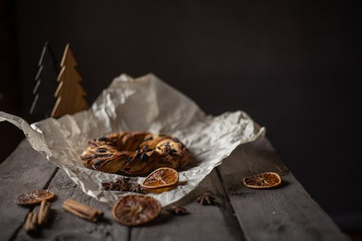 A rustic cinnamon pastry on parchment with dried oranges and spices, captured in moody lighting.