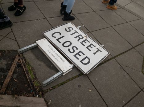A fallen street closed sign on pavement with pedestrians walking nearby.