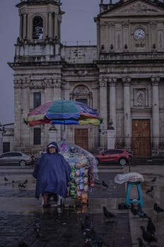 A street vendor under an umbrella sells snacks in front of a cathedral on a rainy day.