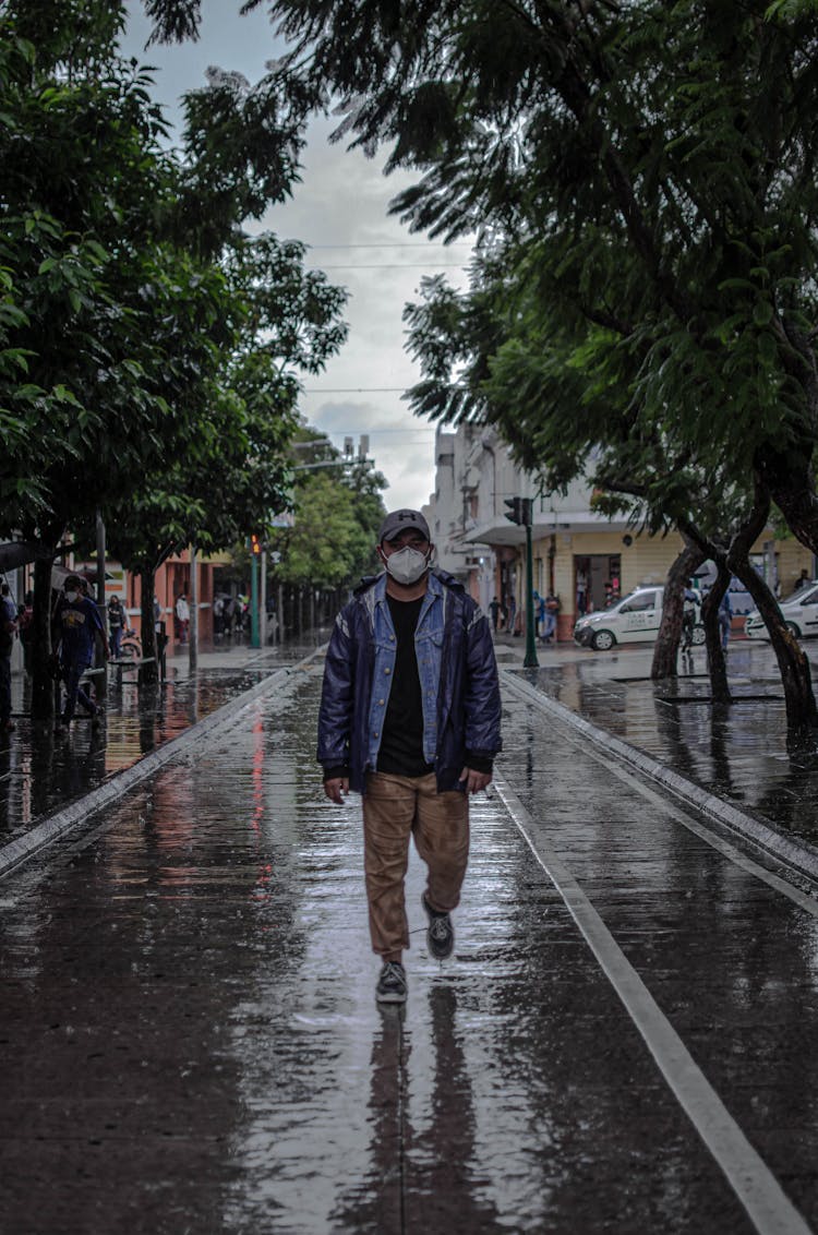 A Man In Mask Walking On The Wet Road With Trees
