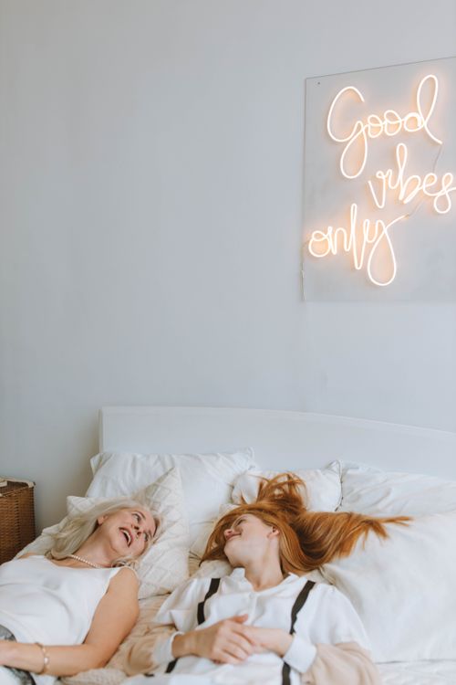 Free Two women lying on a white bed, enjoying a laid-back moment under a 'Good vibes only' neon sign. Stock Photo
