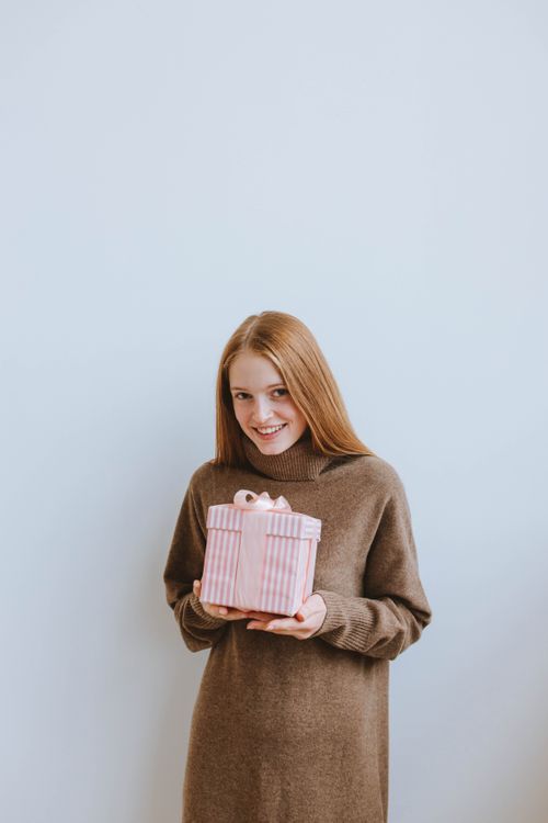Free Smiling woman in brown dress holding a pink-striped gift indoors, perfect for holiday themes. Stock Photo