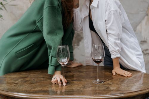 Free Two adults leaning over a wooden table with wine glasses, sharing a secret. Cozy and intimate atmosphere. Stock Photo
