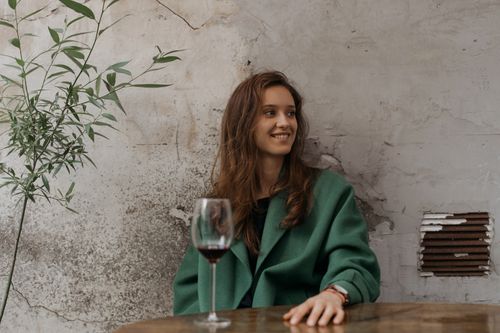 Free A woman sitting outdoors, smiling with a glass of red wine in hand, wearing a green coat. Stock Photo