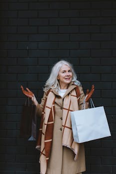 Senior woman with shopping bags against a brick wall, smiling joyfully.