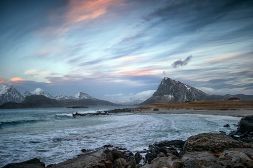 Powerful ocean with foamy waves washing sandy beach surrounded by rocky formations and mountains against cloudy sunset sky in Lofoten Archipelago