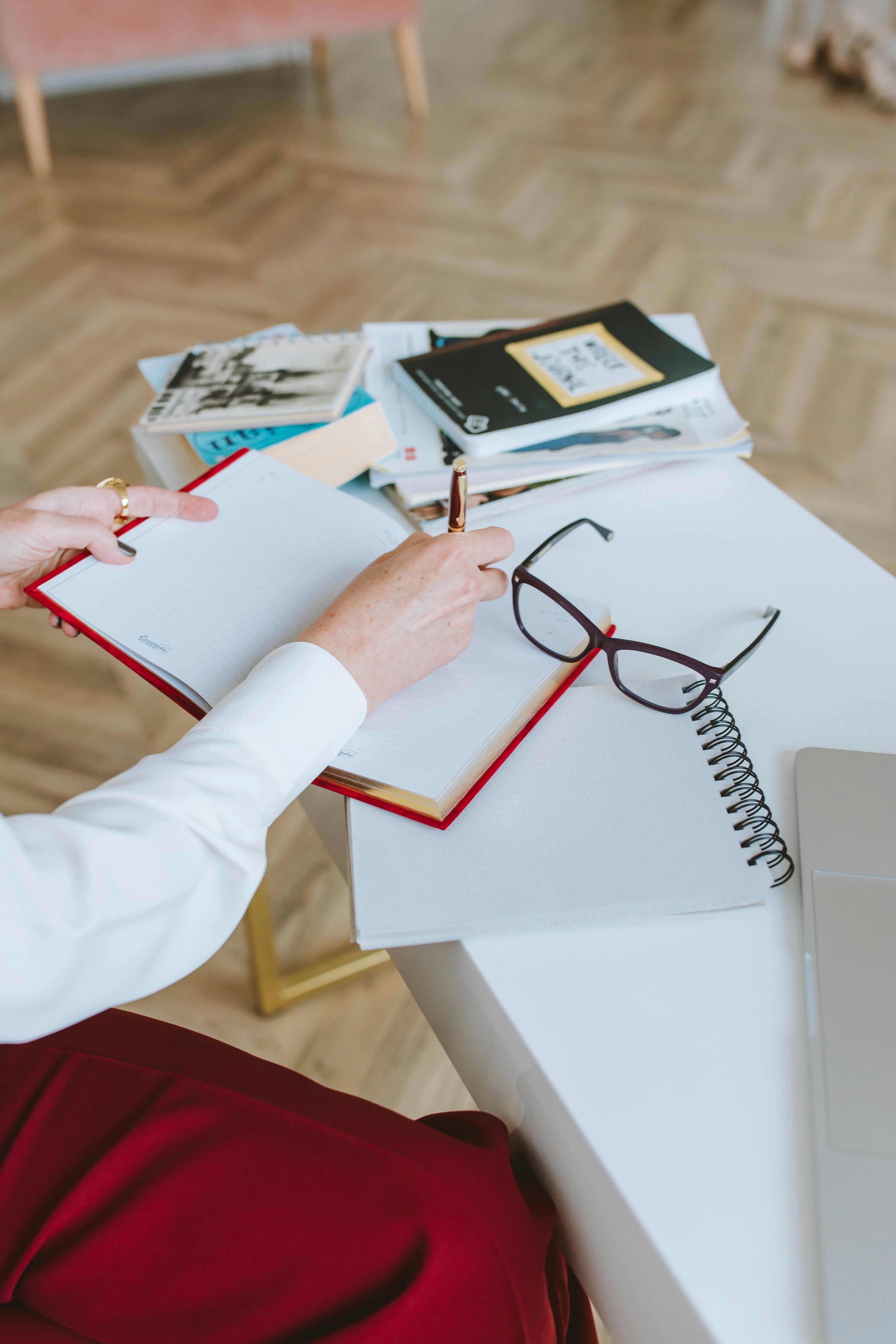 Free Close-up of a person writing in a notebook with glasses and books nearby. Perfect for work or study themes. Stock Photo