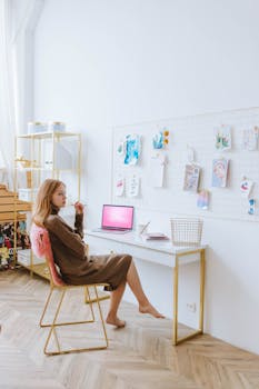 A young woman works remotely in a minimalist home office with a moodboard and laptop.