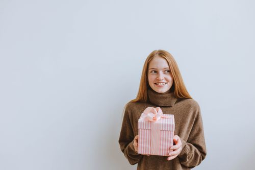 Free A cheerful woman in a cozy sweater holding a pink gift box with a bow, perfect for celebrations. Stock Photo