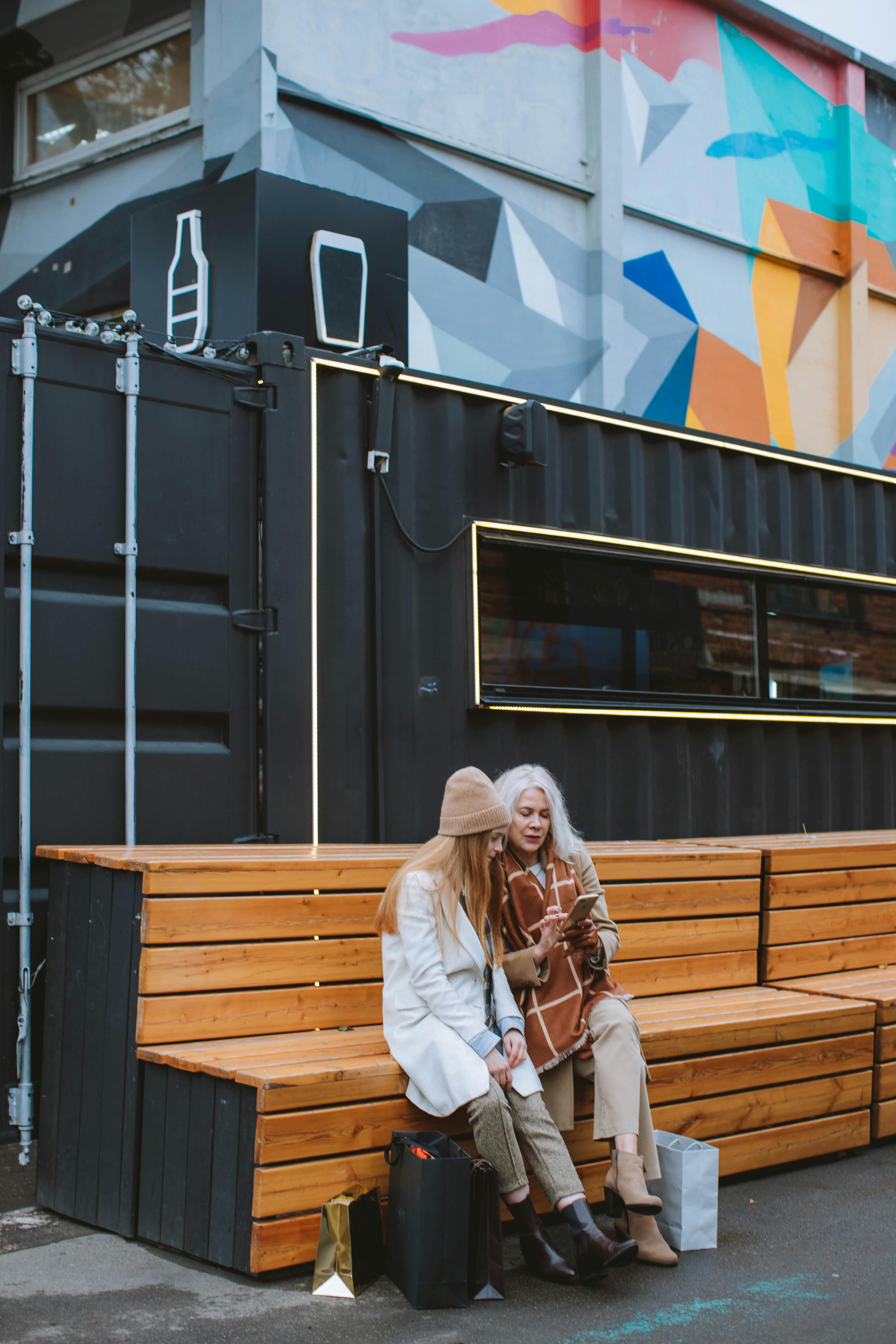 woman with brown scarf and girl in white coat sitting on brown wooden bench and looking at mobile phone beside gray container