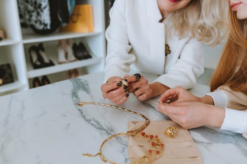 Two Women in White Trying On Golden Rings and Necklaces on Marble Table