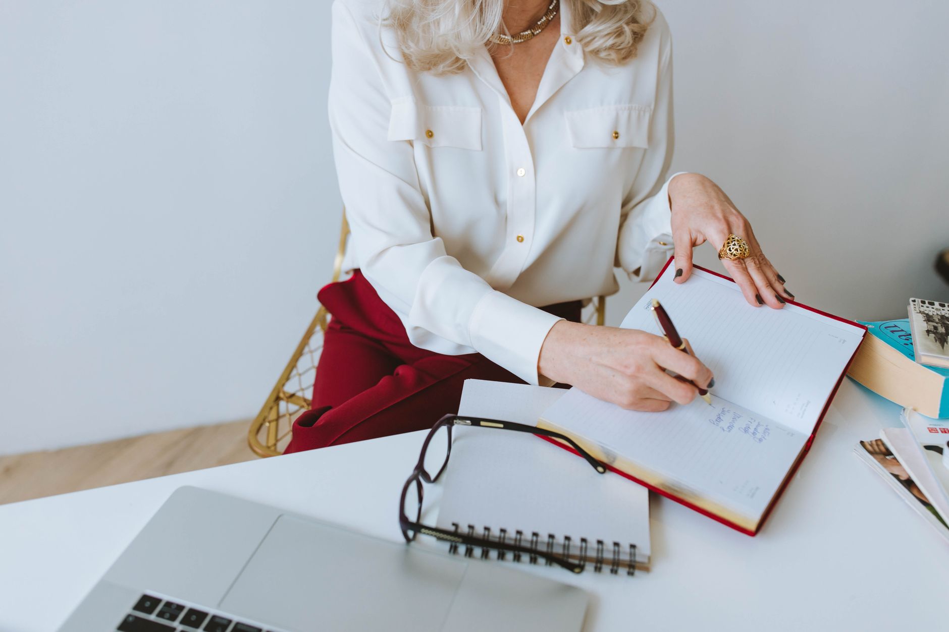 A woman with manicured nails writing in a diary at a white desk with a laptop.