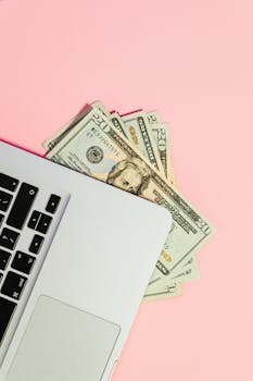 Overhead shot of dollar bills tucked under a laptop on a pink background, symbolizing online transactions.