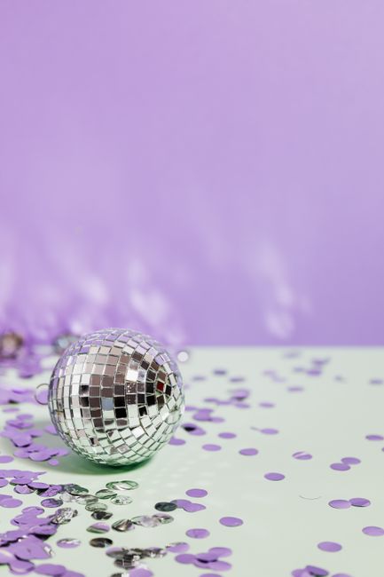 Silver disco ball amid vibrant purple and green confetti with a light purple background