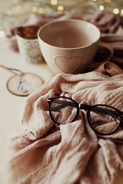 High angle of modern eyeglasses on pink textile near empty ceramic cup and lights at home
