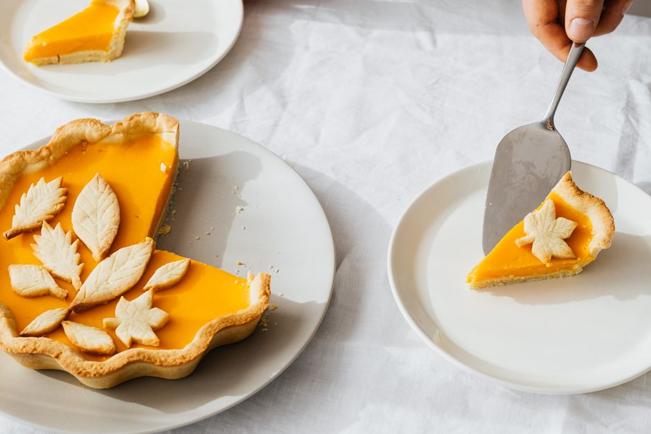 A slice of pumpkin pie on a white plate with a dollop of whipped cream, showing the smooth orange filling and flaky crust edge, with the whole pie in the background.