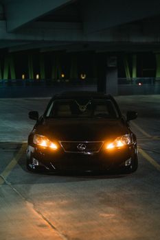 Front view of a black Lexus parked in a dimly lit garage at night, headlights on.