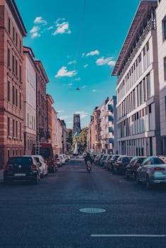 Motorcyclist driving on empty paved roadway with cars on sides among low rise aged buildings under blue sky