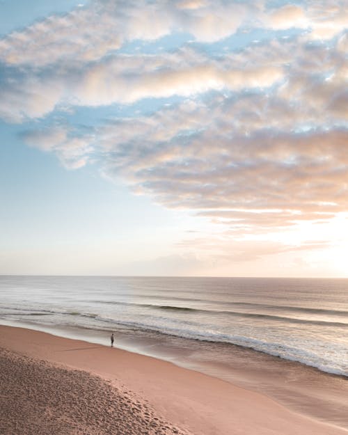 Calm anonymous person standing on sandy beach and admiring wavy ocean under colorful sky at sunset