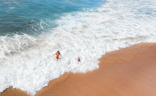 Drone view of unrecognizable female traveler in bikini walking in foamy ocean near sandy beach during summer holidays