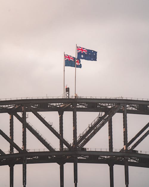 Scenery of national Australian flags waving on majestic Sydney Harbor Bridge arch trusses under gray sky at dusk