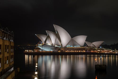 Glossy white facade of majestic Sydney Opera House in expressionism style reflecting in calm river water at dark night