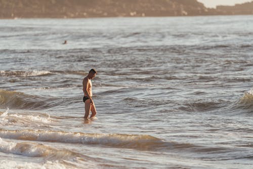 Side view calm fit male in black swimming trunks standing in shallow waving seawater on early evening