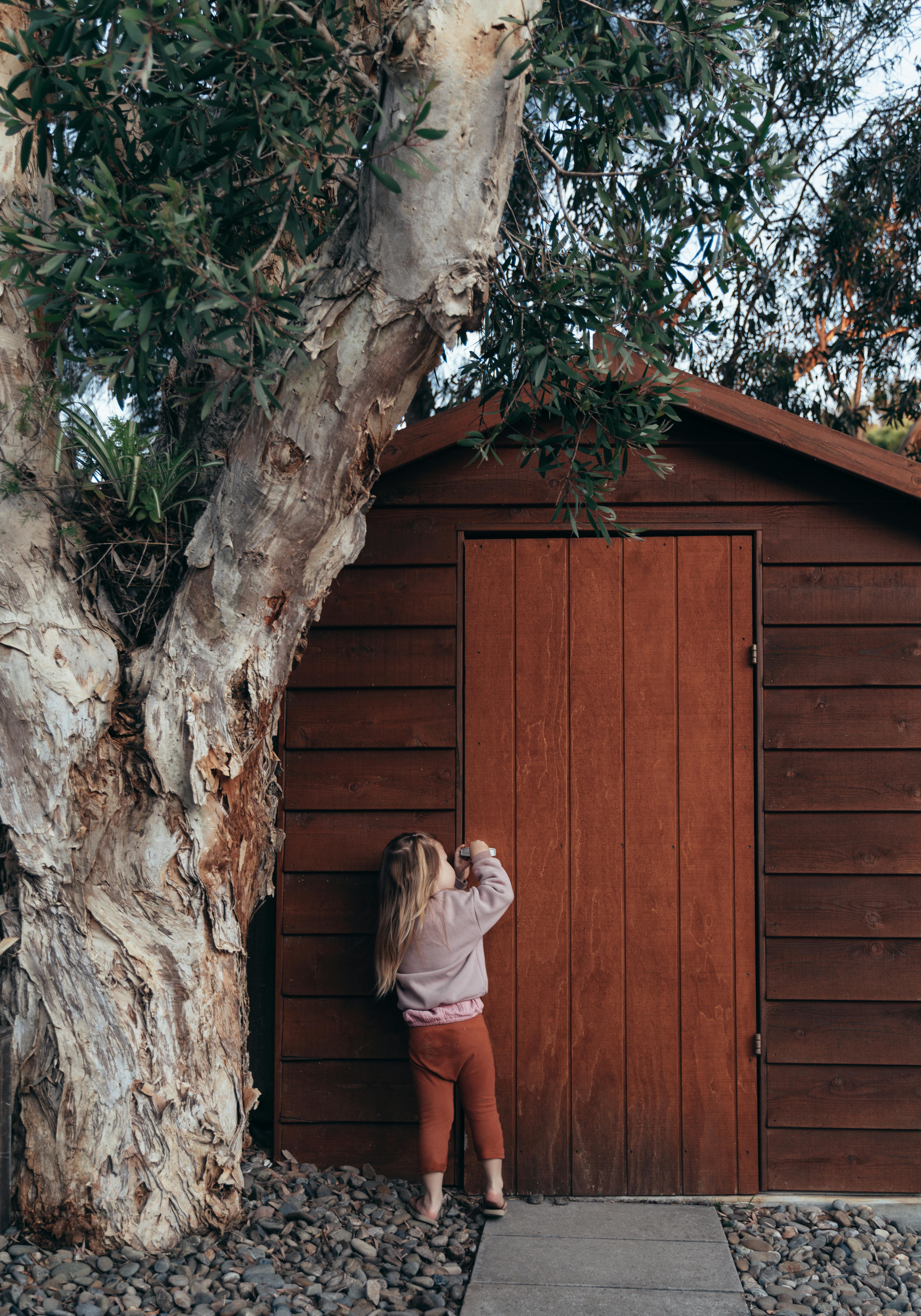 girl opening door of wooden house near tree