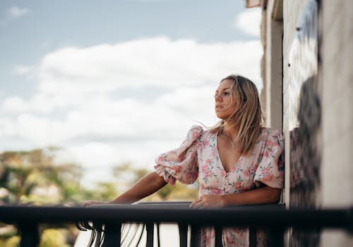 Young content serious female in delicate dress looking away and leaning on railing of terrace