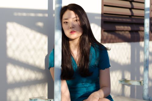 Free Young Asian woman in blue top leaning on metal pipe and looking at camera in sunlight Stock Photo