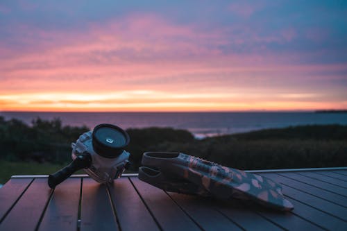 Action camera placed near flippers on wooden table in nature on seaside against grassy lawn and sunset sky over sea with blurred background