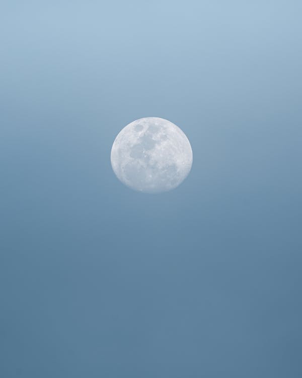 Full white round moon with craters on surface shining on cloudless clear sky in nature on summer day high in air