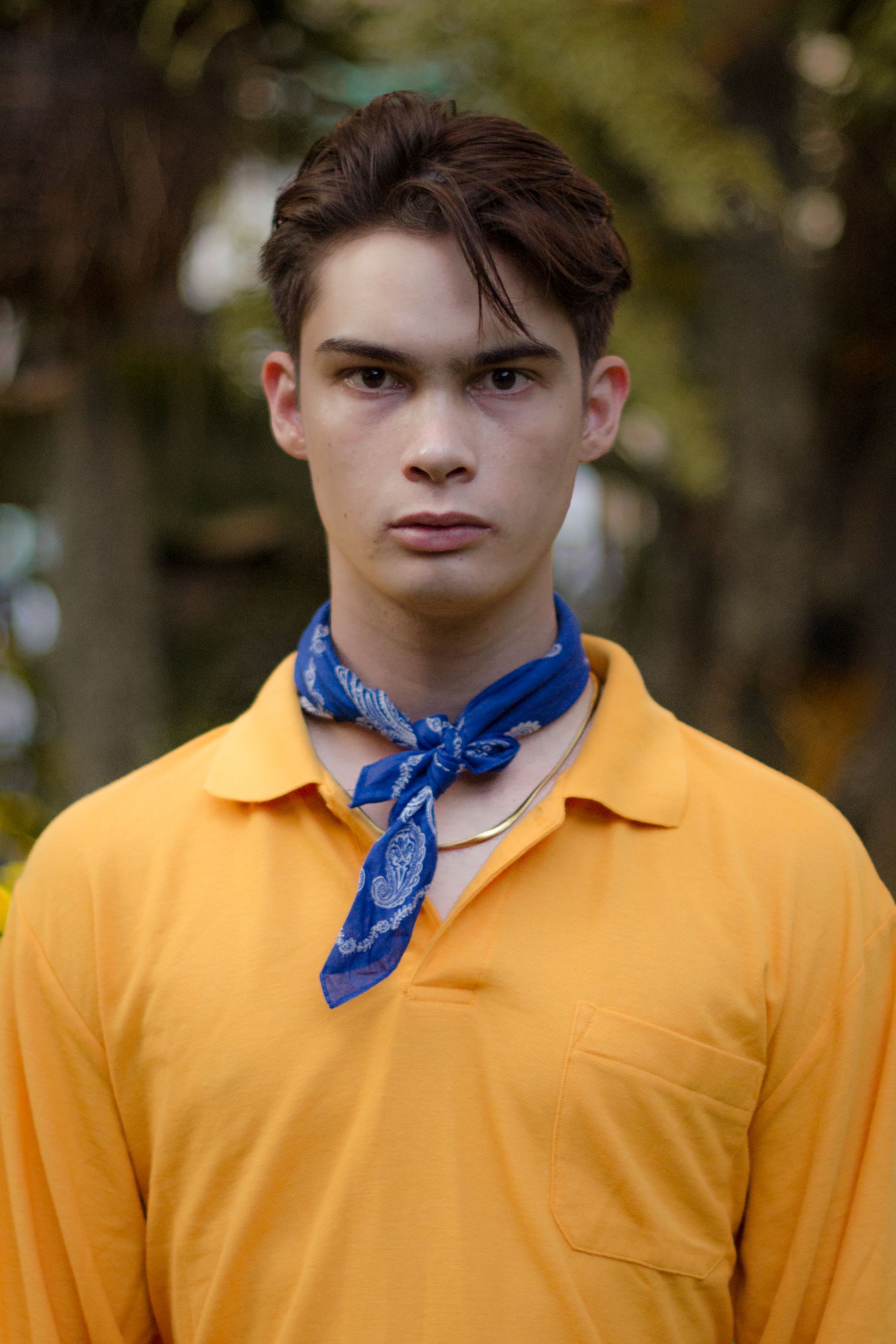 Young handsome guy wearing orange shirt and blue handkerchief looking seriously at camera on blurred background